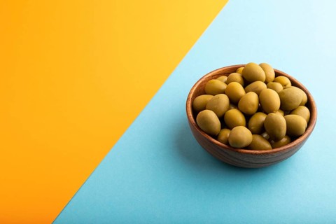 Fresh green olives in wooden bowl on blue and orange pastel paper background. side view, copy space