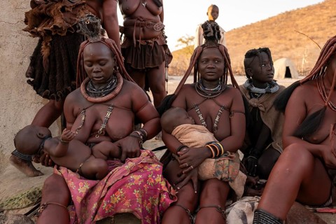 Married Himba woman with their babies in their arms sitting in front of the first woman's hut, in