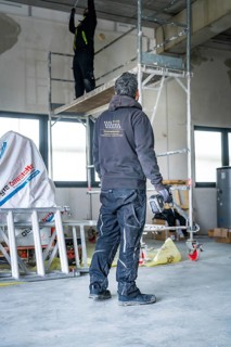 A worker on scaffolding works in a construction site, another worker standing next door,