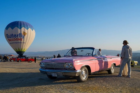 Person standing next to pink cabriolet and hot air balloon under clear fresh morning sky,