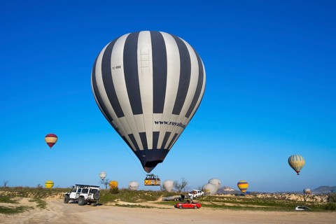 Large striped hot air balloon lands next to jeep and red car in clear landscape, Göreme Tarihî