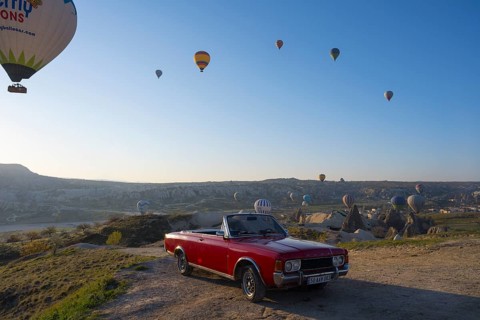 A red convertible stands in front of a vast landscape with hot air balloons in the morning sky,