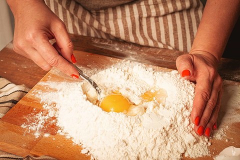 A person with bright nails is combining eggs with flour on a wooden board. The setting is a cozy
