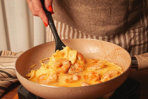 A person stirs shrimp and eggs in a frying pan, showcasing a home-cooked meal. The cozy kitchen