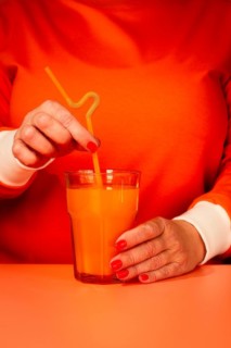 A woman in an orange sweater, stirring with a straw, orange juice in a glass, orange background