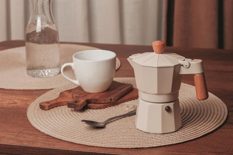A moka pot sits on a wooden cutting board beside a white cup on a round placemat. A glass bottle of
