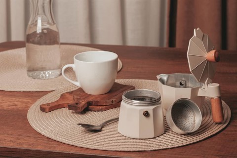 A cozy coffee setup features a white cup, a moka pot, a spoon, and a water bottle on a wooden table