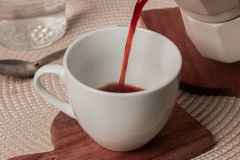 Hot coffee is poured into a white cup resting on a wooden coaster. A serene breakfast setting is