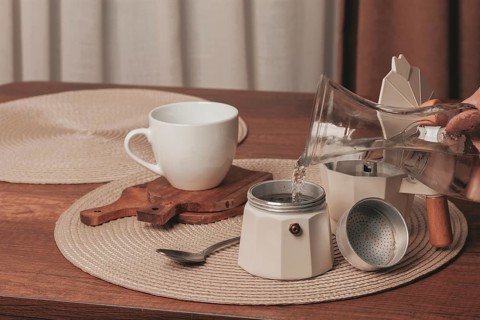 Hot water is being poured from a glass kettle into a coffee maker on a wooden table. A white cup