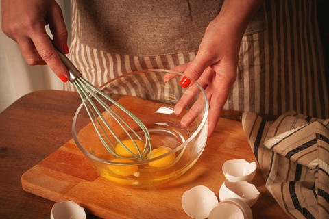 Hands are mixing fresh eggs in a glass bowl with a whisk. Egg shells are scattered on the wooden