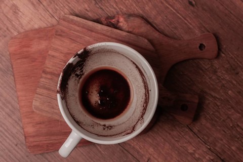 A warm coffee cup sits on a wooden cutting board, showing traces of dark liquid and coffee grounds.