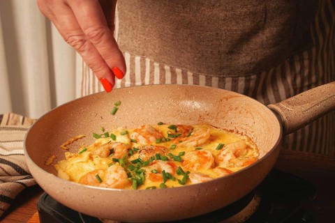A woman is cooking shrimp in a skillet, adding fresh herbs to the dish. The warm colors of the