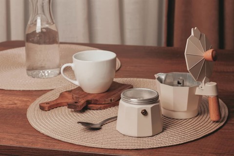 A cozy scene features a white cup resting on a wooden coaster next to a moka pot and a glass water