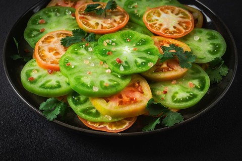 Sliced green tomatoes, on a dark surface, garnished with herbs and salt, top view