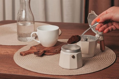 A hand holds a spoonful of coffee grounds over a moka pot while a cup and a bottle of water sit on
