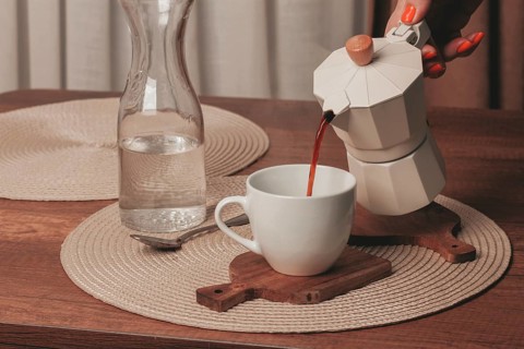 A hand pours freshly brewed coffee from a stovetop maker into a white cup. A glass pitcher filled