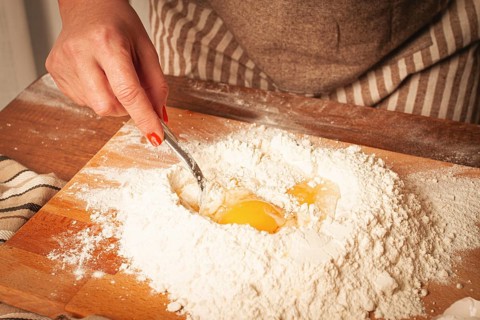 A hand with bright nails mixes eggs into a pile of flour on a wooden board. The cozy kitchen