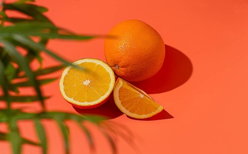 On the coral surface, next to two neatly sliced slices, there is a whole orange. Sunlight casts