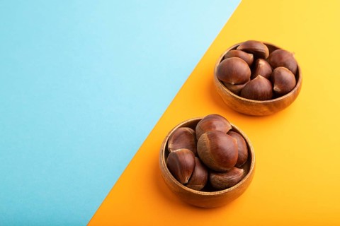 Wooden bowl with raw edible Ð¡hestnuts on blue and orange paper pastel background, side view, copy