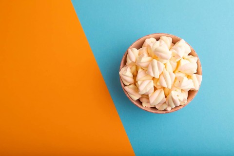 Orange and pink marshmallow in ceramic bowl on blue and orange pastel paper background, top view,