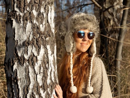 Slender pretty woman with fur hat and pompoms, red hair and warm winter colors between birch forest
