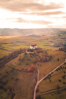 A red church roof on a hill with a view of vast fields under a cloud cover, Wurmlinger Chapel,