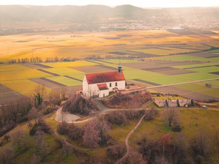 A church with a red roof towers over a vast mosaic of fields in sunlight, Wurmlingen Chapel,