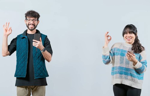 Smiling couple holding a cell phone and gesturing OK, isolated. Happy young couple holding phone
