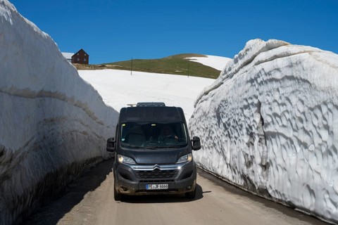 A van drives through thick snow walls on a mountain road under a blue sky, motorhome, camper on a