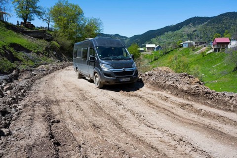 A van drives down a muddy road surrounded by green hills and individual houses, campers on the way