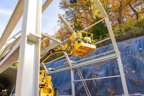 Workers in a yellow lift work on a raised metal structure in front of a forest, Hermann Hesse