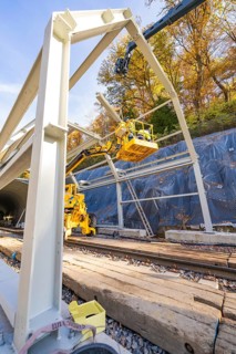 Construction workers erect a metal structure on rails along a wooded area, Hermann Hesse Railway