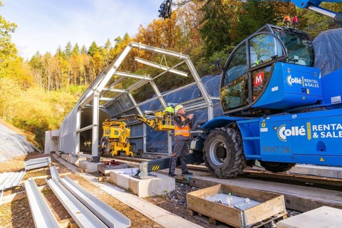 Workers use cranes to build a structure along a rail line in the forest, Hermann Hesse Railway bat
