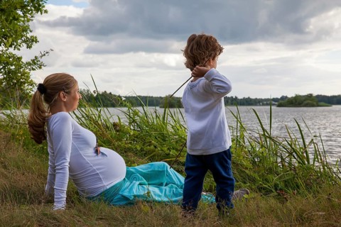 Pregnant woman with little boy at the lake, in grass, child playing with sticks