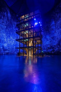 An illuminated, multi-storey structure in front of a blue artificial forest projection, Amazonia