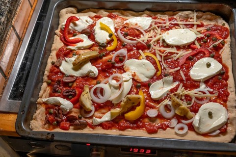 Homemade pizza topped with gorgonzola and tomatoes, olives and peppers on a baking tray in front of