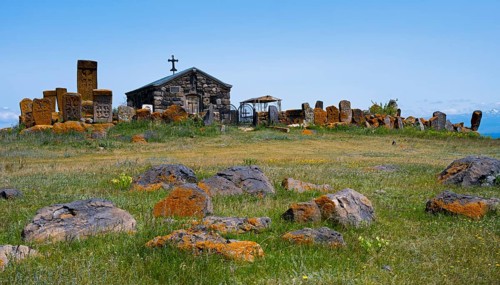 Small chapel surrounded by old tombstones on a hilly meadow under a blue sky, khachkar at the