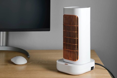 A sleek electric heater stands on a wooden desk next to a computer monitor. The heater features a