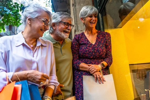 Senior friends laughing together while window shopping on a city street, carrying multiple shopping
