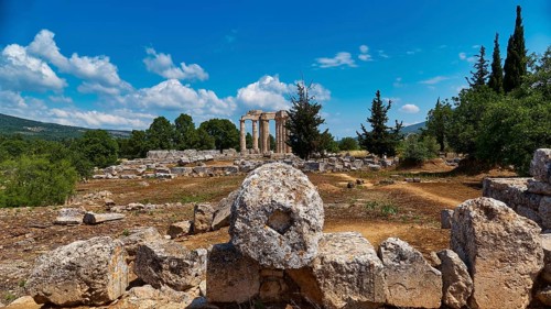 Ancient temple columns in an open field with surrounding trees and mountains in the background,