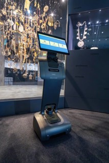 A robot with a screen stands in front of a glass display case with decorative objects in a museum,
