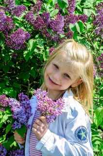 Little blonde 7-year-old girl stands surrounded by blooming lilacs and smiles at the camera in