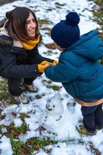 Happy mother and young son build a small snowman outdoors, laughing and bonding in warm parkas,