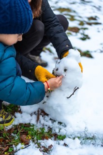 Young boy and mother collaborating to build a small snowman, carefully adding details like eyes and