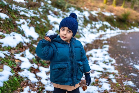 Young boy in warm parka and hat holding a snowball, smiling while playing in a snowy forest