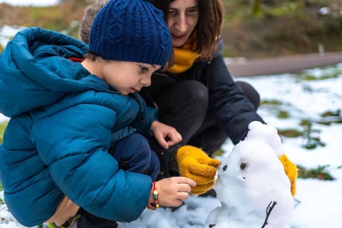 Mother and young son crouch in fresh snow, building and adding details to a tiny snowman while