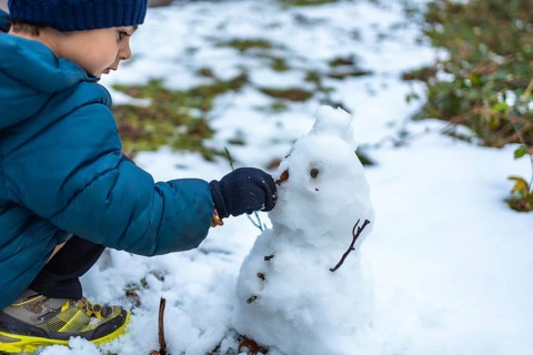 Young boy playing in the snow, carefully placing a small natural element on a snowman he is