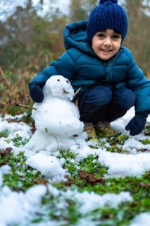 Smiling young boy crouching outdoors, happily creating a small snowman on a patch of grass lightly