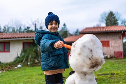Smiling little boy in warm winter clothes builds a snowman in the yard, holding a carrot to place