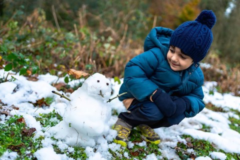 Young boy kneeling on snowy ground, wearing a warm coat, hat, and gloves, observing a freshly made